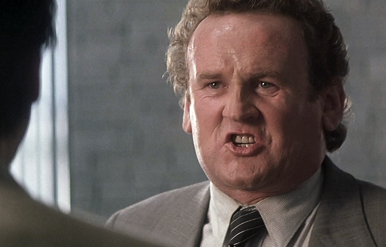 Närbild på en arg, lätt svettig man i grå kostym och randig slips som skriker med blottade tänder mot en person i förgrunden, i en scen ur Con Air.