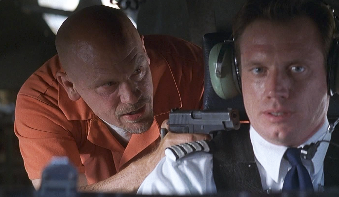 I en scen ur Con Air håller en flintskallig fånge i orange overall en pistol mot en pilot med headset och uniform i cockpit.