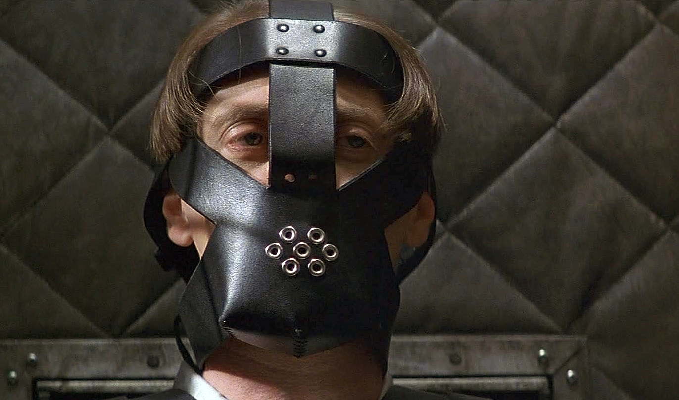 Man med page och allvarlig blick bär en svart lädermask med nitar och små metallringar över ansiktet, framför en vadderad vägg. Scen ur Con Air.