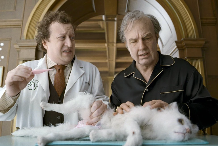 En veterinär i vit rock och en man i svart pyjamas med guldkant står över en vit katt som ligger utsträckt på ett bord, båda med bekymrade miner.