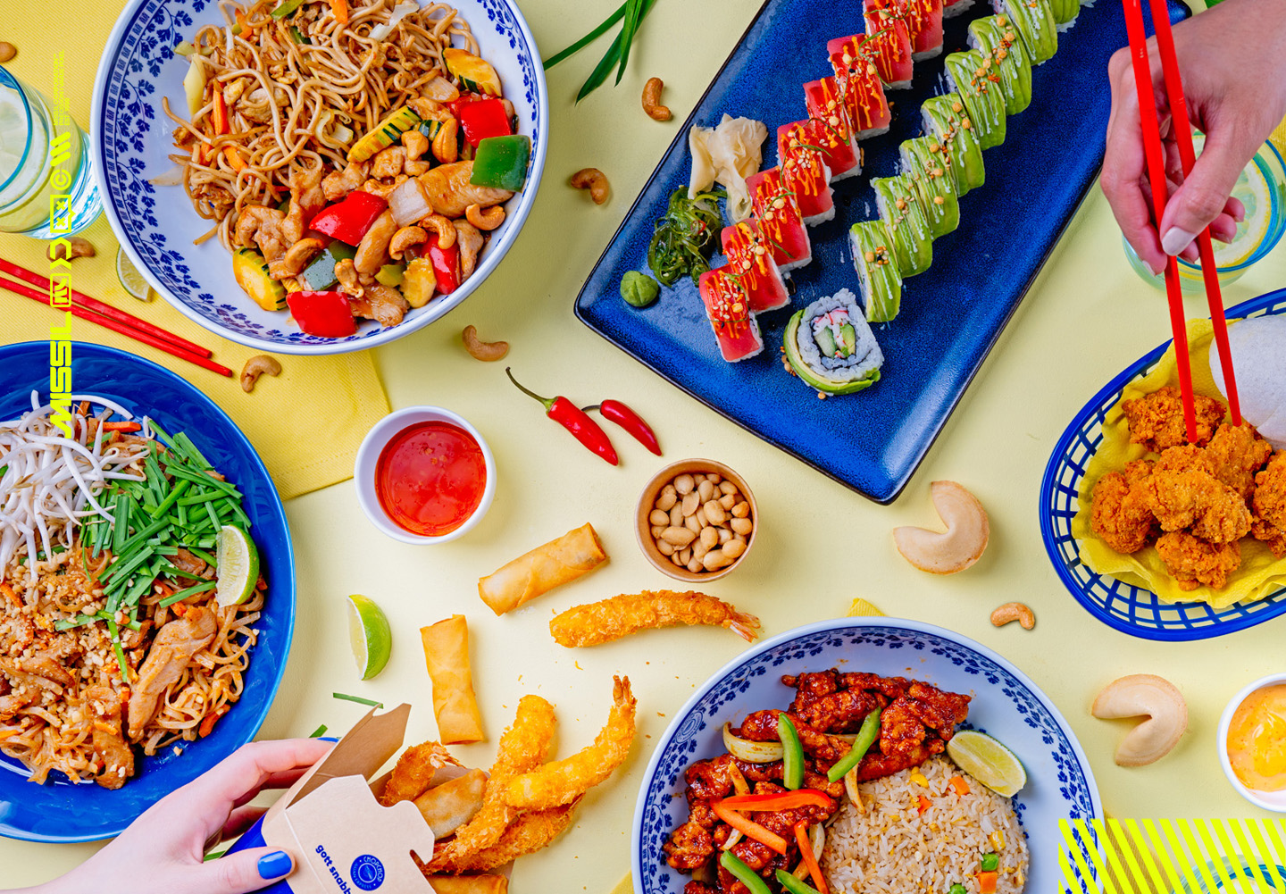Asiatiskt festbord sett uppifrån med nudlar, sushirullar, friterade räkor, vårrullar, kyckling och stekt ris på blå tallrikar, samt ätpinnar och lyckokakor.