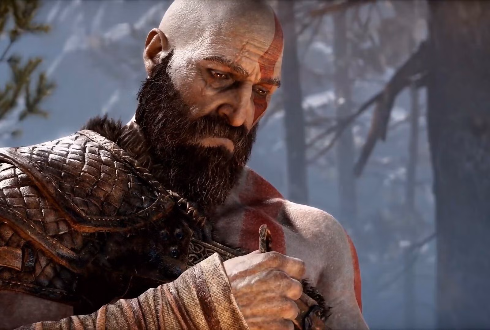 Kratos med flintskalle, mörkt skägg och röd ansiktsmålning blickar nedåt allvarligt i en snöig skog, klädd i en fjällig läderrustning.