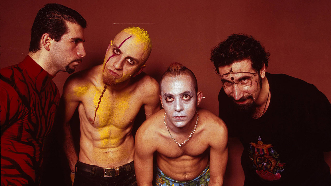 Fyra män i bandet System of a Down poserar mot en rödbrun bakgrund. Två är barbröstade med gul respektive vit ansiktsmålning, de andra två bär röd mönstrad tröja och svart t-shirt.
