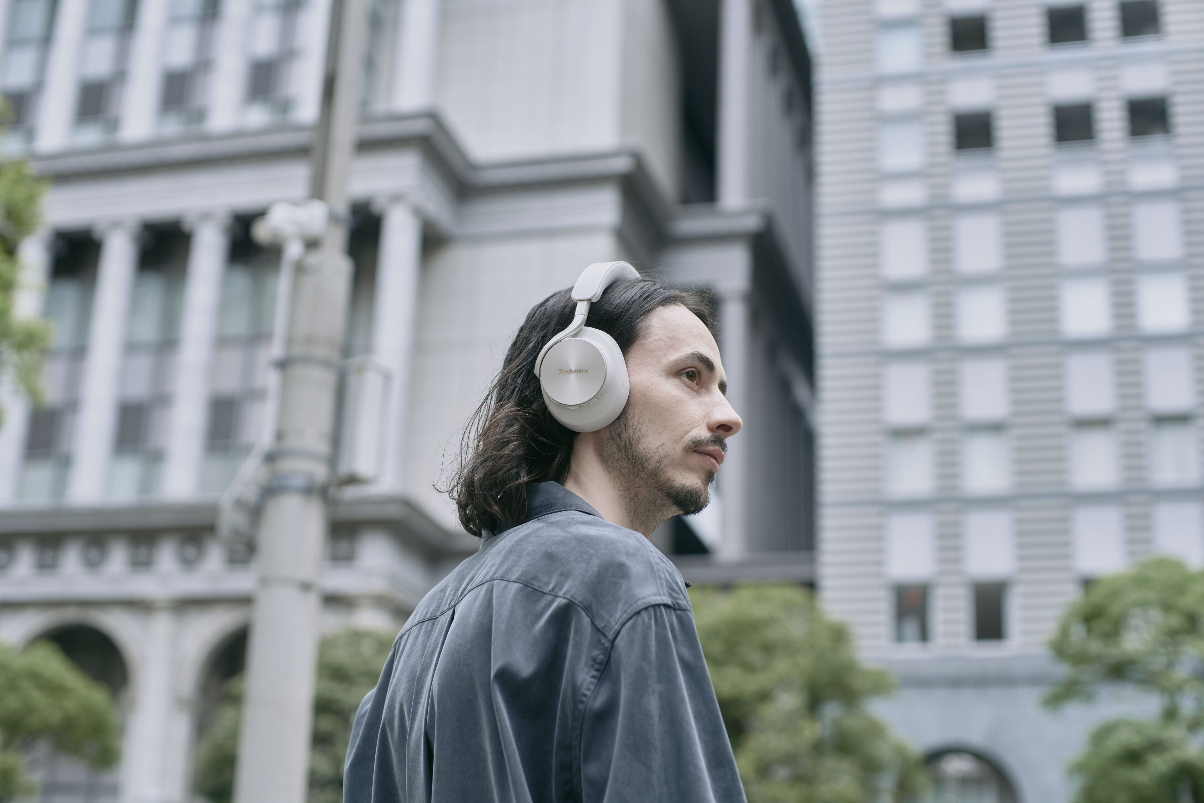 Man med långt mörkt hår och skägg tittar åt sidan på en stadsgata, iklädd ett par ljusbeiga hörlurar av over-ear-modell från Technics, med stora byggnader i bakgrunden.