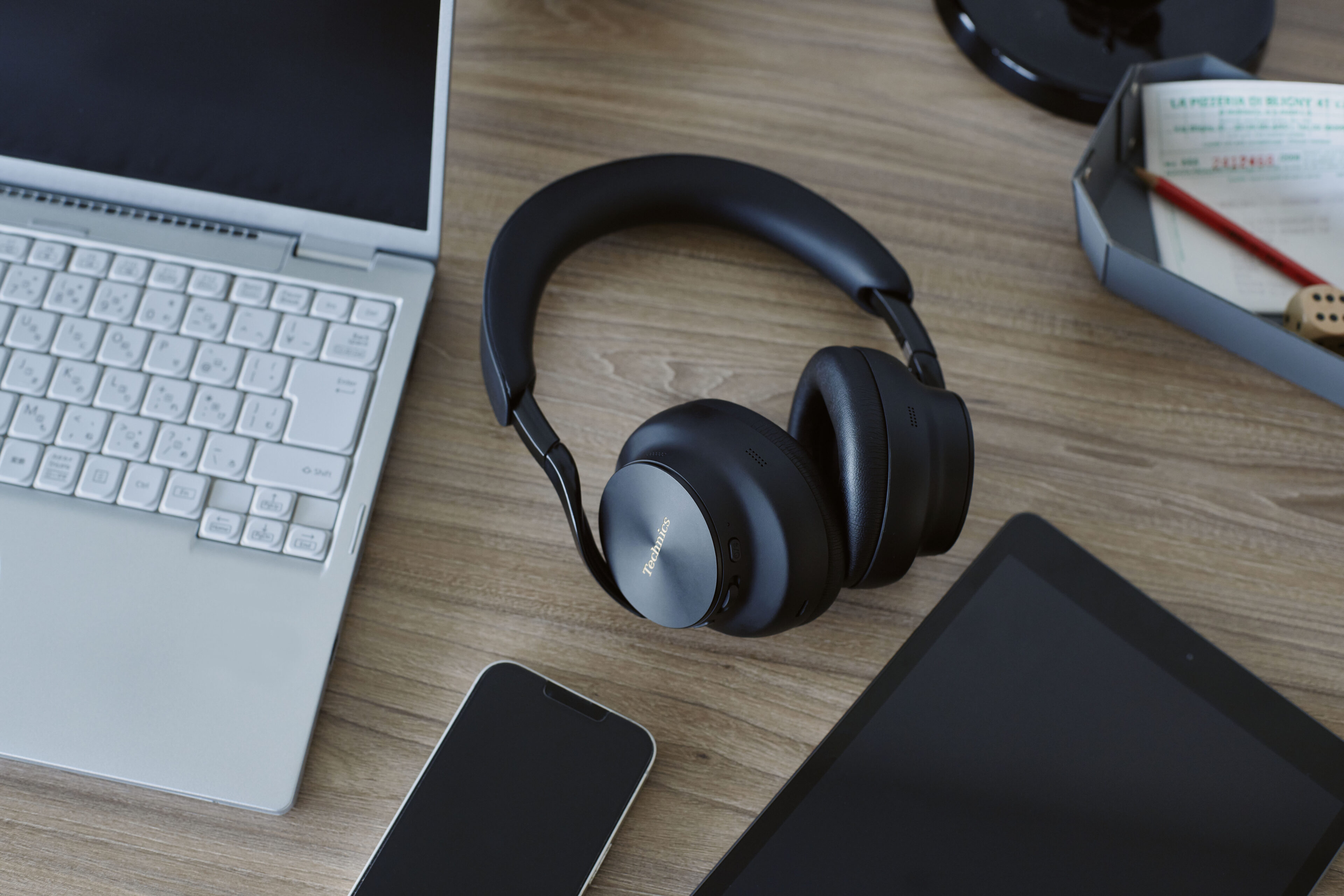 Svarta over-ear-hörlurar från Technics ligger på ett träskrivbord mellan en silverfärgad laptop, en mobil och en surfplatta.