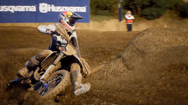 Motocrossförare i leriga kläder lägger sin motorcykel i en tvär sväng på en lerig bana i MXGP 26, med en stor jordkant till höger och en funktionär i bakgrunden.