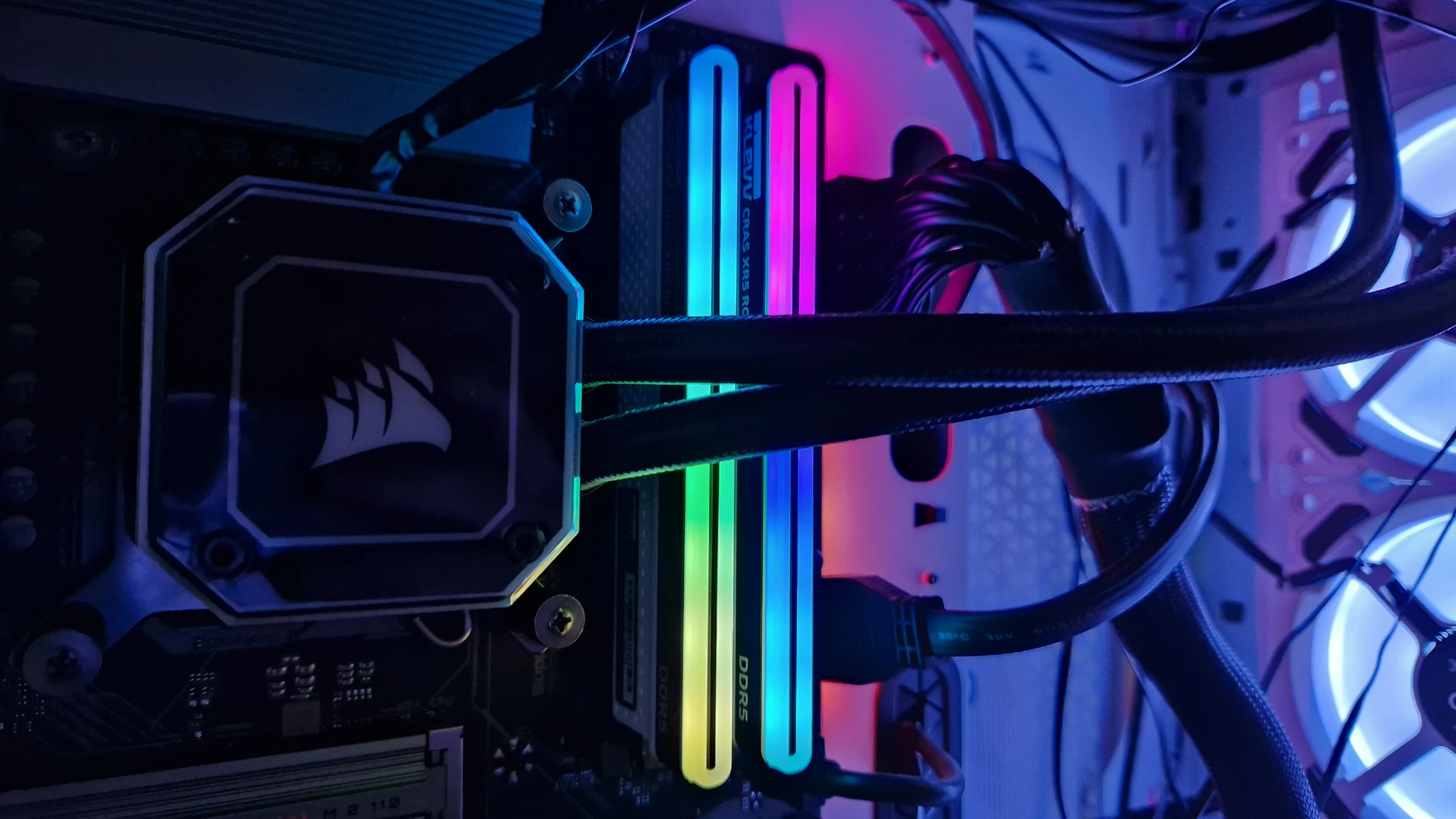 Närbild av det inre i en spel-PC med Corsairs vätskekylning och lysande RGB-minnesmoduler av typen DDR5 i regnbågsfärger, omgivna av flätade kablar och lysande fläktar.