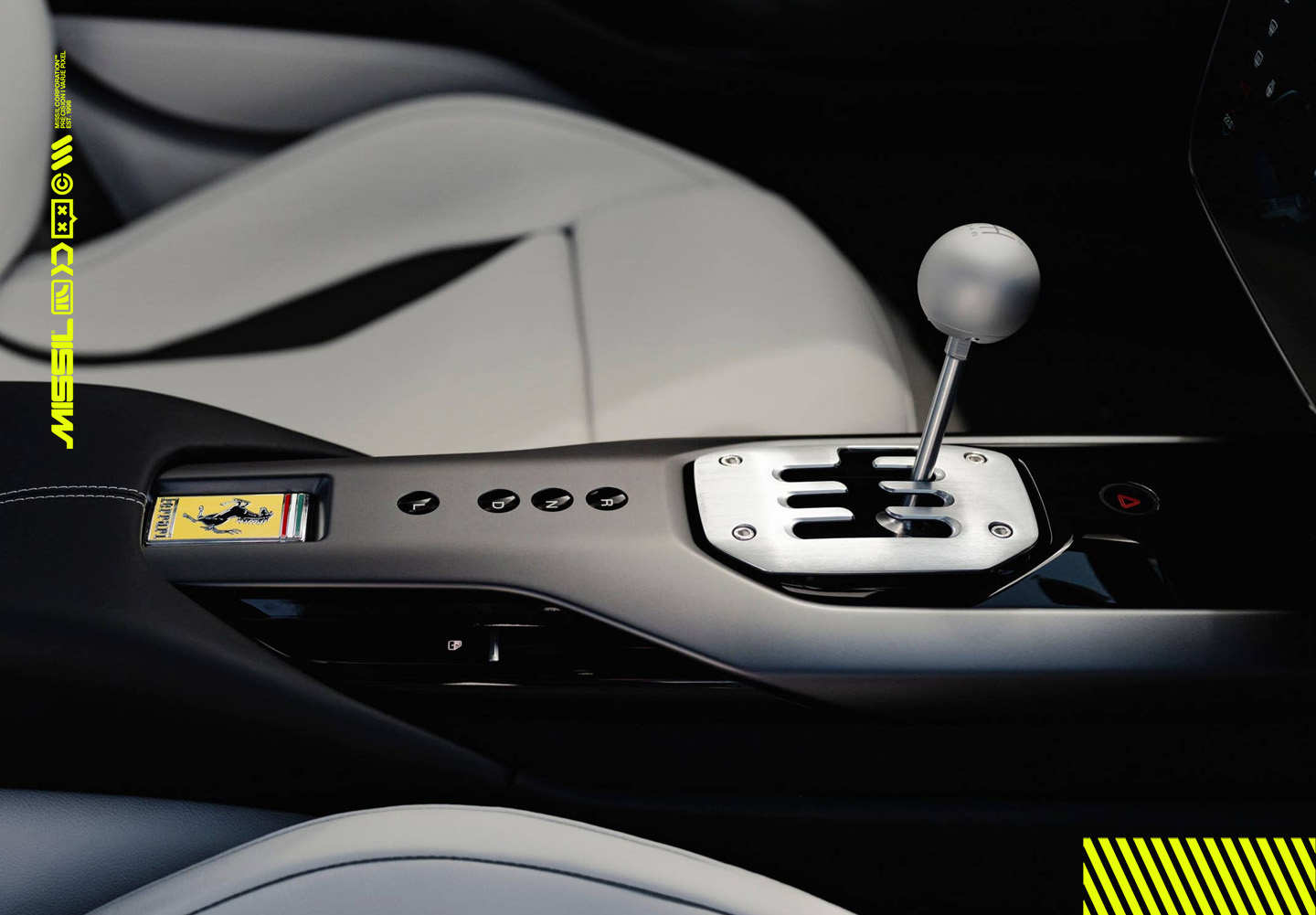 Närbild på växelspaken i en Ferrari med silverfärgad knopp och öppen växelkulissen i metall, mellan ljusgrå lädersäten. Ferraris hästemblem syns på mittkonsolen.